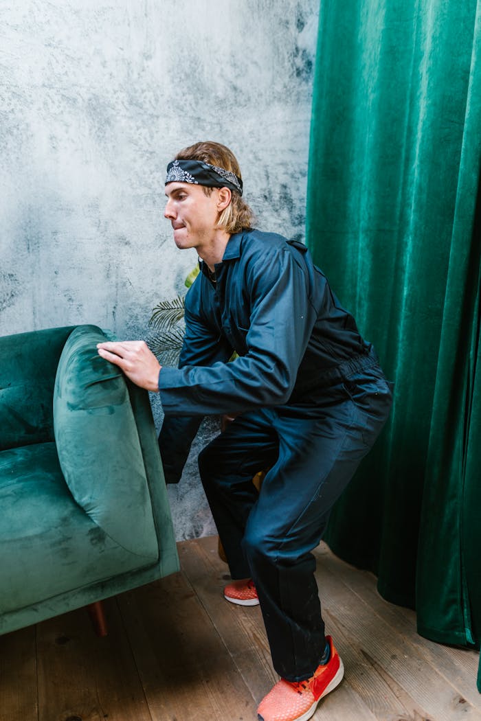 A man in a bandana and casual clothing lifting a green couch indoors.